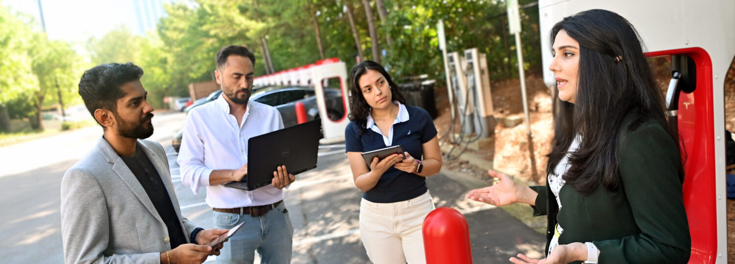 Leila Hajibabai talking with three students while standing in front of an electric car charging station.